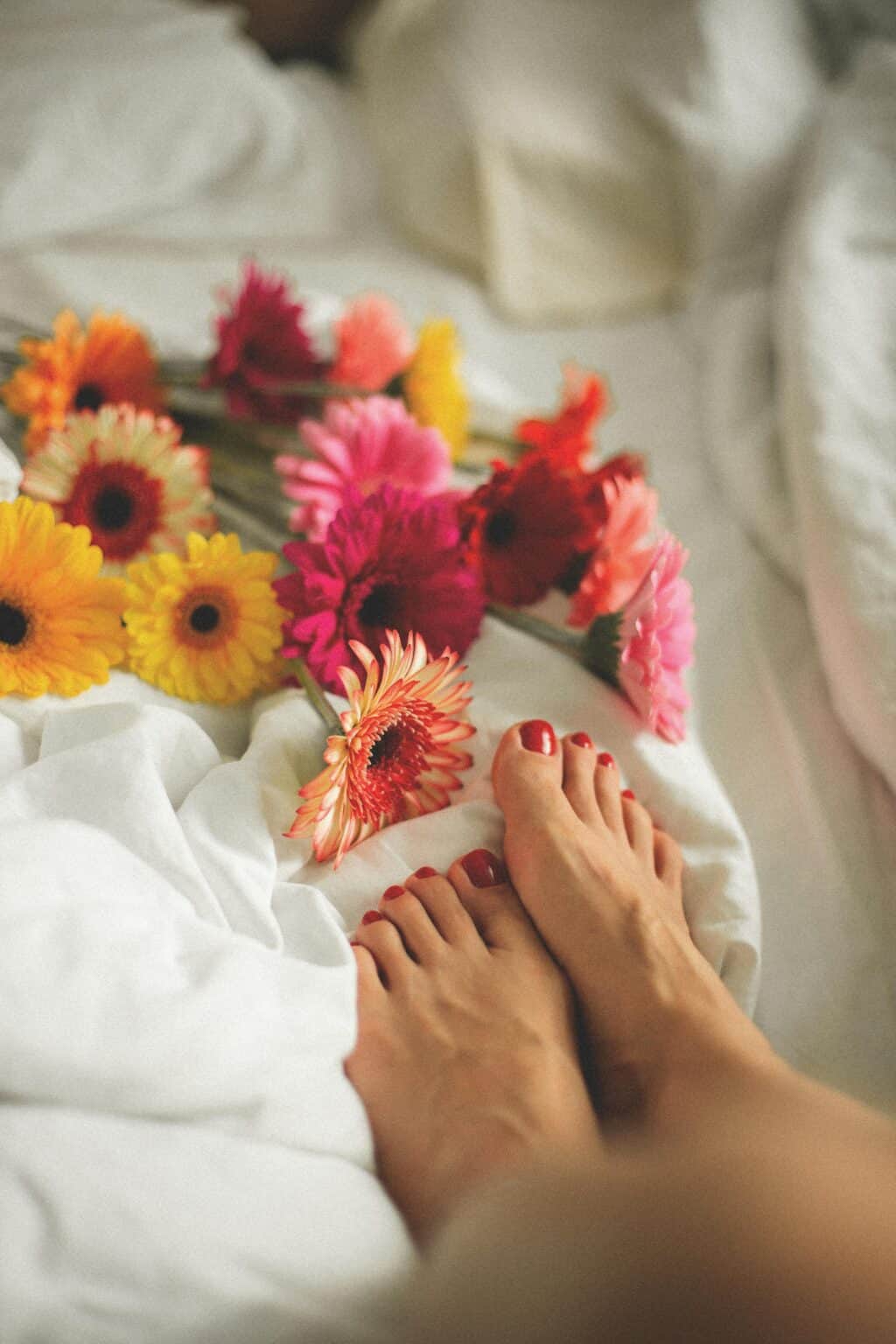 Colorful gerbera flowers and woman's feet on a white bed create a serene and delicate setting.